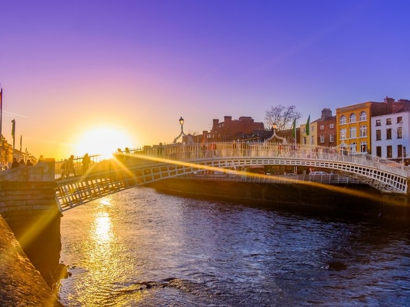 Sunset over a river in Dublin, with the Ha'penny Bridge silhouetted against a vibrant sky. The soft glow reflects off the water, creating a serene atmosphere.