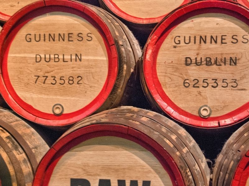 Stacked wooden barrels with red rims, labeled "Guinness Dublin" and numbers. The image conveys a rustic, historical feel, suggesting an old brewery.
