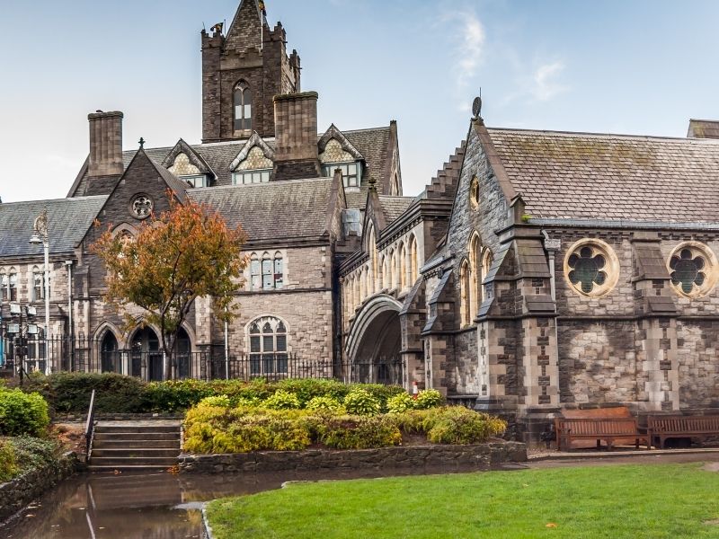 Historic stone church with intricate Gothic architecture, arches, and round windows. Surrounding lush garden and benches create a peaceful ambiance.