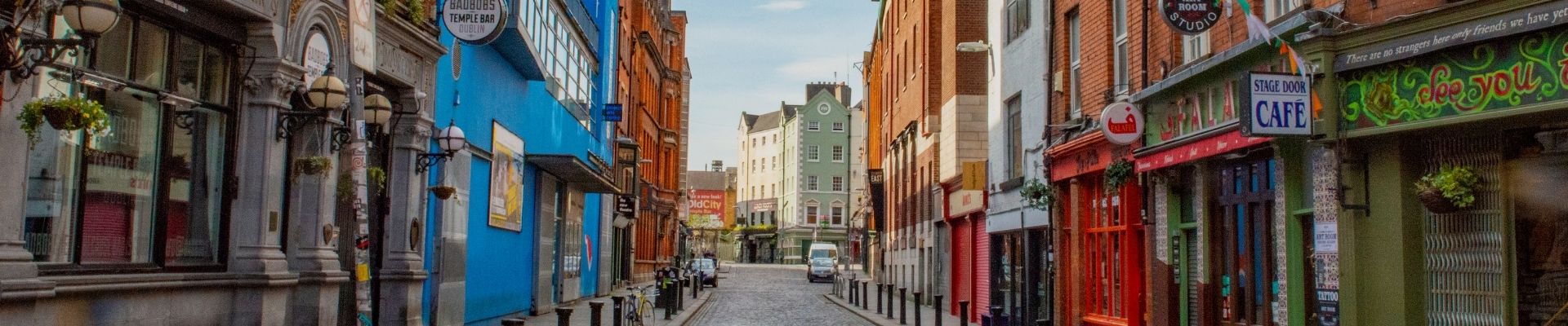Narrow street in an urban area with colorful buildings on both sides; a vibrant blue wall and red-bricked facades. Bright signage and shops convey a lively atmosphere.