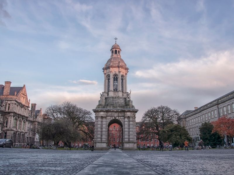 A grand stone bell tower stands in the center of a cobblestone courtyard, flanked by historic university buildings and trees. A few people walk and cycle through the open square under a soft, cloudy sky.