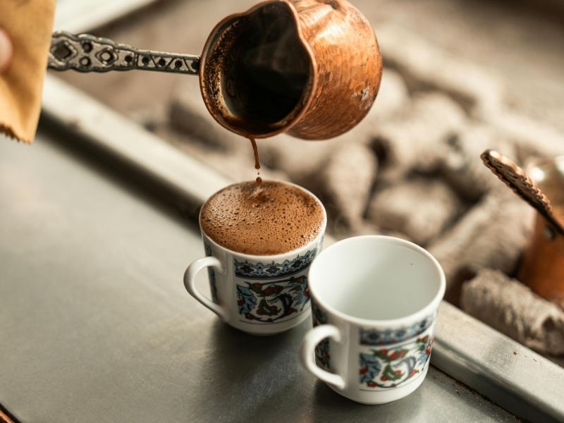 A copper pot pours frothy Turkish coffee into an ornate, patterned cup beside an empty matching cup.