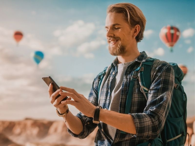 young handsome tourist hiking with rucksack using smartphone on top a rocky canyon valley at a hot air balloon festival